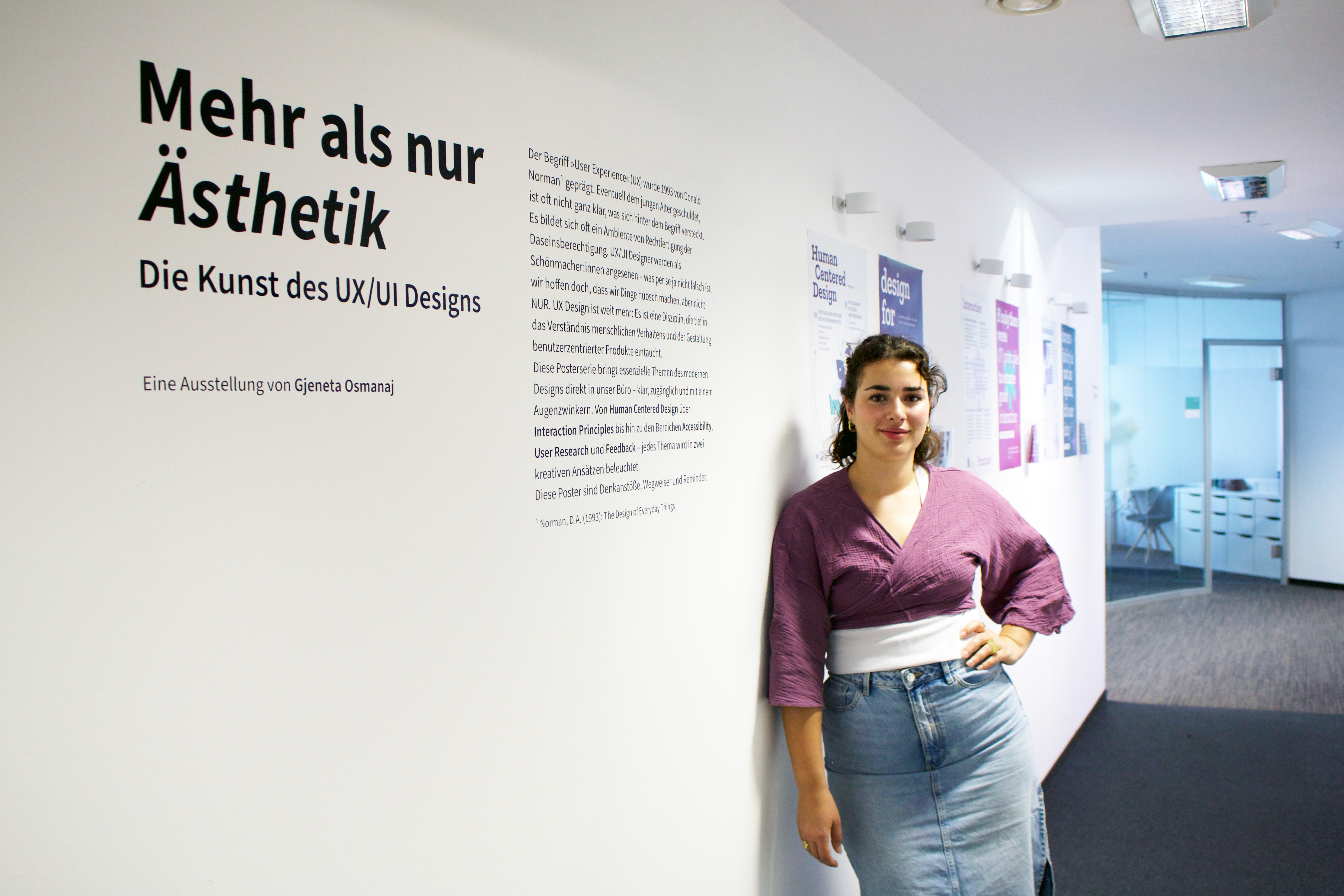Gjeneta Osmanaj stands in a hallway next to a wall displaying the title of her exhibition "Mehr als nur Ästhetik – Die Kunst des UX/UI Designs" ("More Than Just Aesthetics – The Art of UX/UI Design"). She is wearing a purple top and a denim skirt, smiling confidently in front of a series of posters about UX principles and design concepts.
