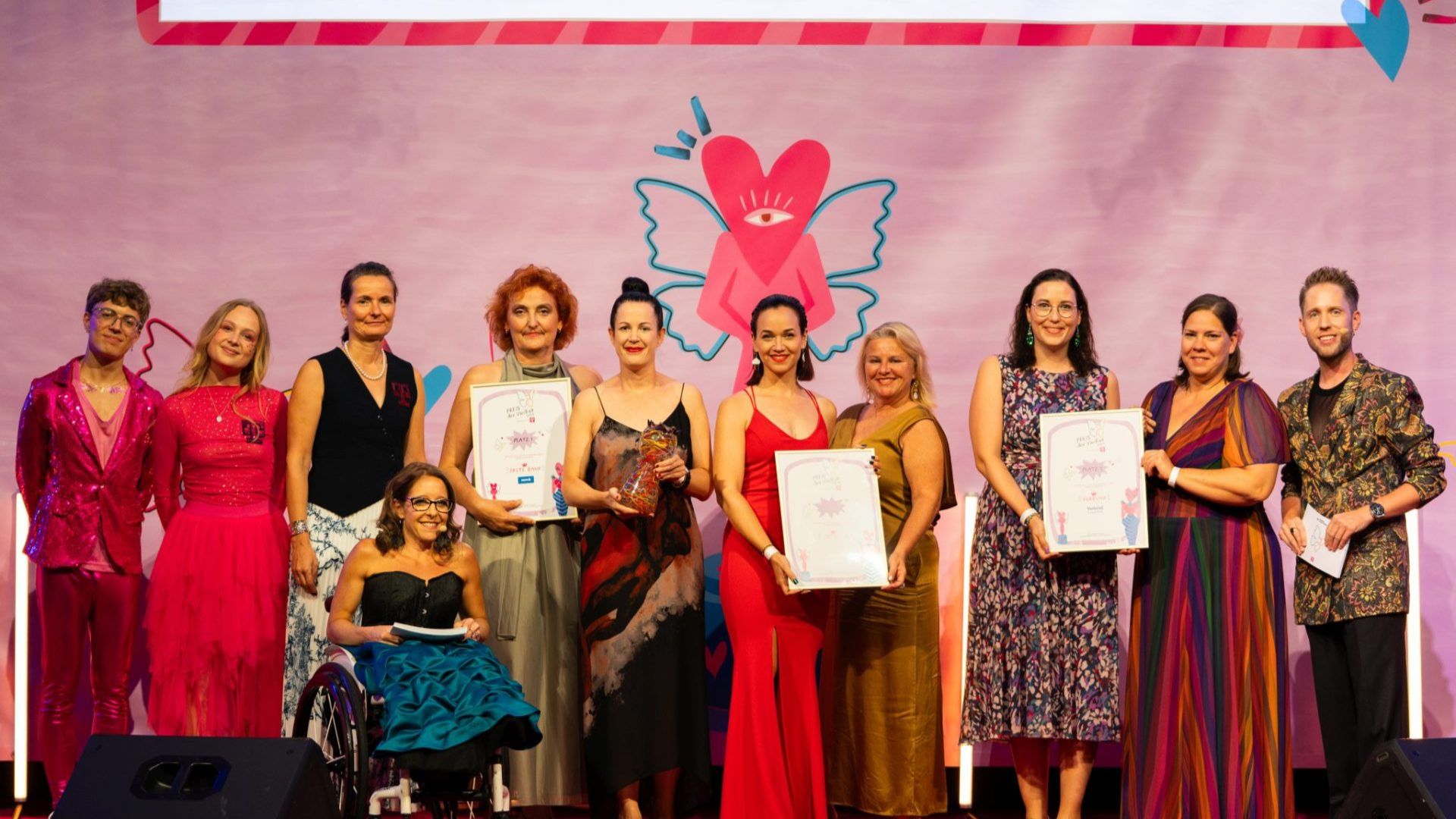 Several women in elegant dresses, one woman in the foreground in a wheelchair, and on the far right a man also dressed formally. Some people are holding picture frames with certificates from an award ceremony.