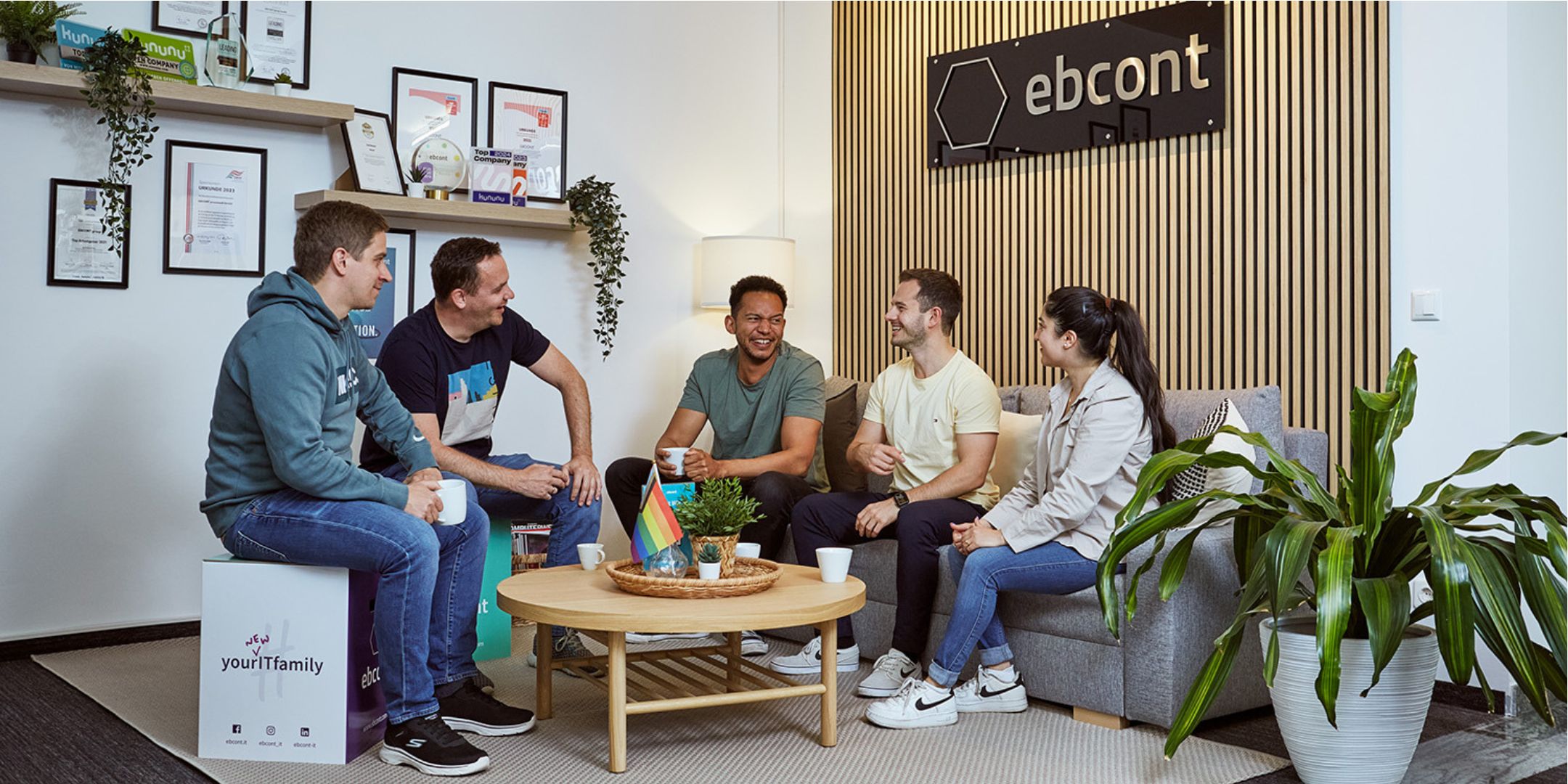 Several people are sitting together on a sofa and chatting. Behind them is a brown wooden wall with the EBCONT logo.