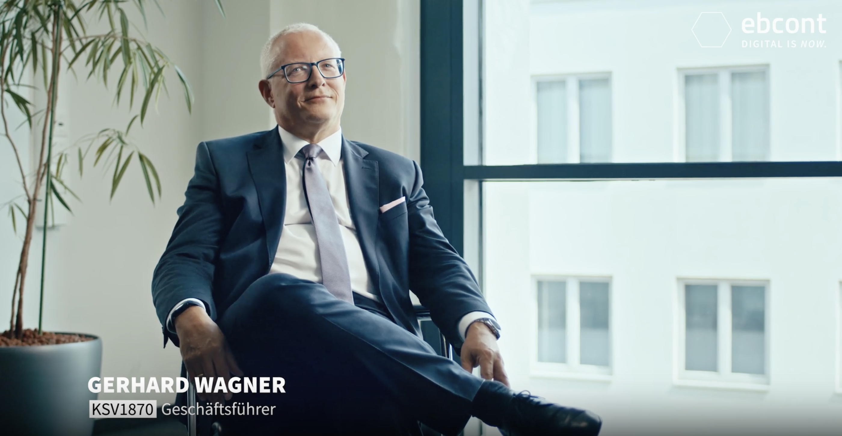 Ein Ausschnitt aus dem Video auf dem Gerhard Wagner entspannt in einem lichtdurchfluteten Raum sitzt und leicht an der Kamera vorbei blickt. Er trägt einen dunkelblauen Anzug mit Krawatte.